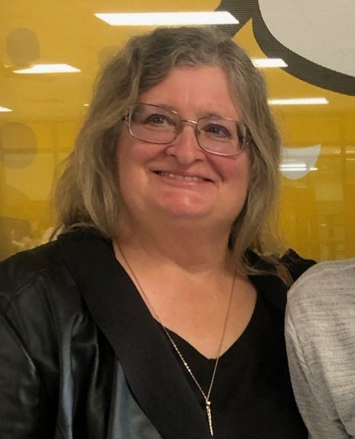 Person wearing a black shirt and necklace standing indoors with a yellow background.